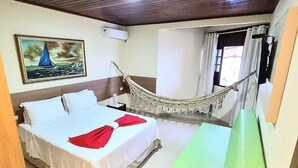 Una habitación de hotel con una cama, una hamaca, un banco verde, un cuadro de un velero y una ventana con cortinas.