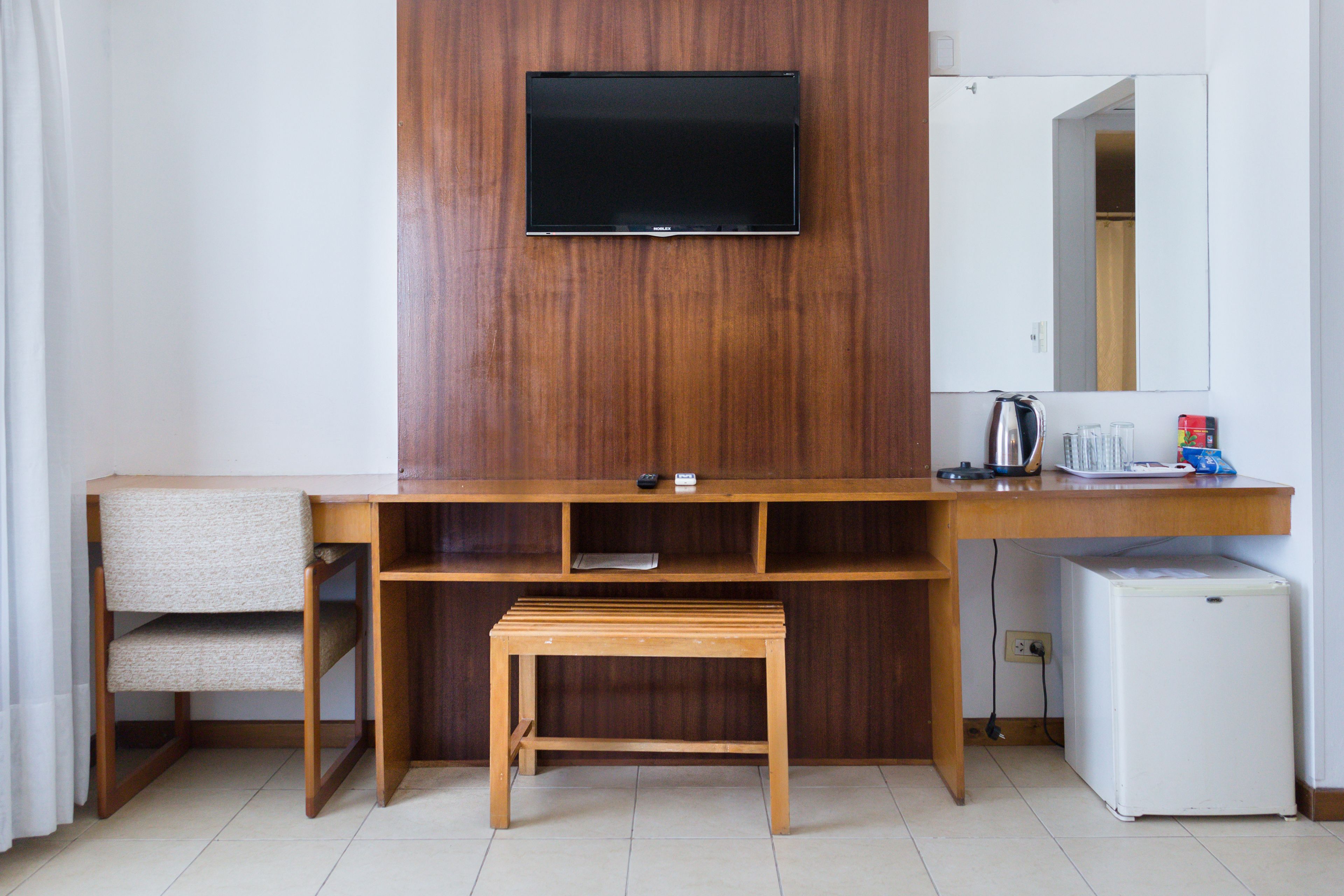 A compact hotel room with a wooden desk, a television mounted on the wall, a small bench, a refrigerator, and a mirror.