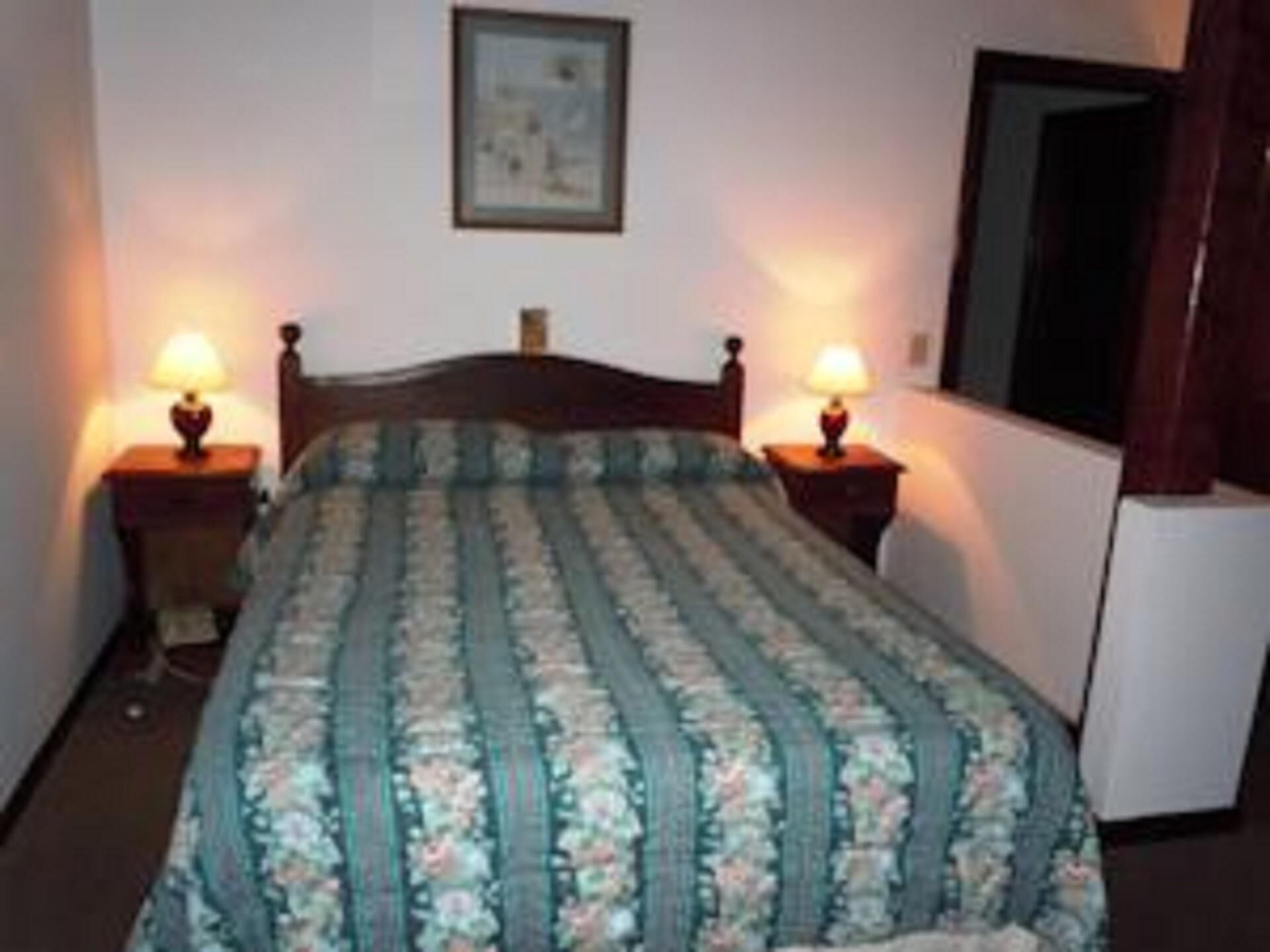Una habitación con una cama, dos mesitas de noche con lámparas, un cuadro en la pared y una puerta a la derecha.