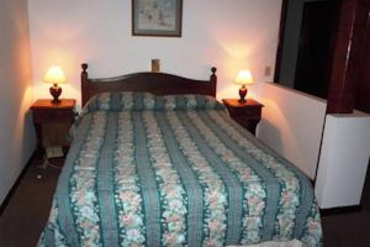 Una habitación con una cama, dos mesitas de noche con lámparas, un cuadro en la pared y una puerta a la derecha.