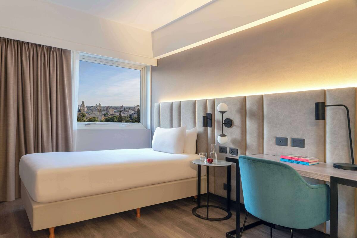 Ein Hotelzimmer mit Bett, Schreibtisch, Stuhl, Lampe und Blick auf eine Stadtlandschaft.