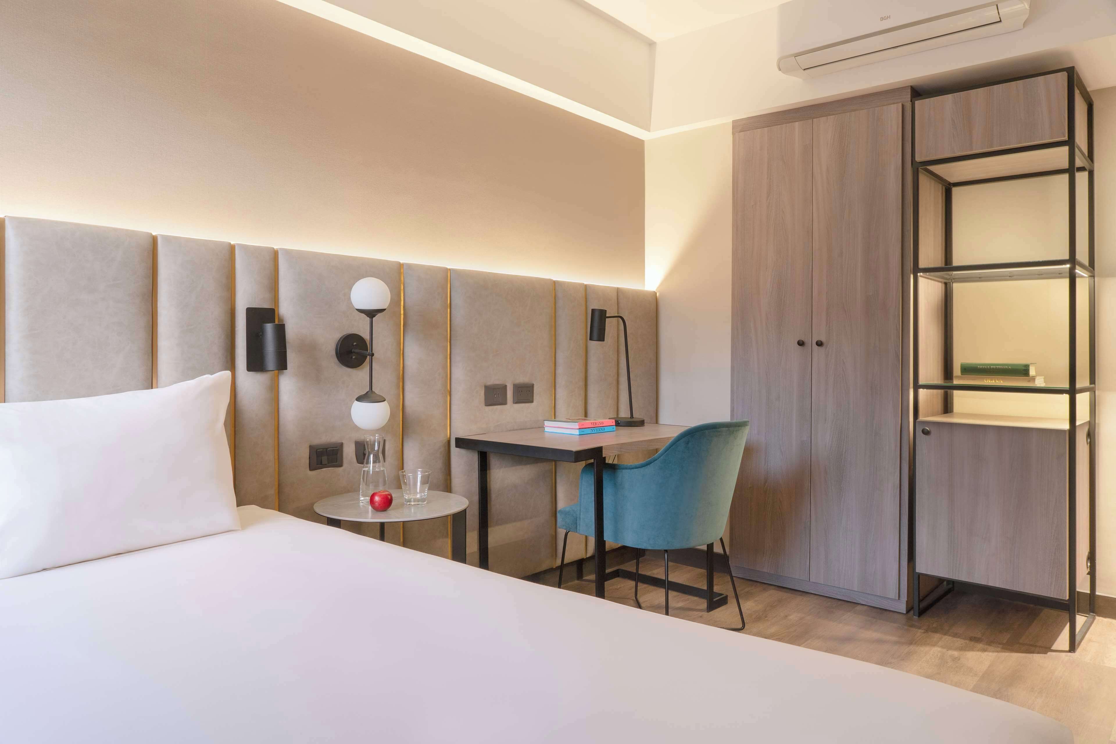 A hotel room with a bed, a desk with a chair, a wardrobe, and a wall-mounted lamp.