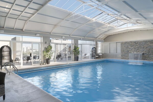An indoor swimming pool with a glass ceiling, stone wall, and outdoor seating area.