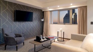 A modern hotel room with a flat-screen TV mounted on the wall, a ceiling fan, a large window with curtains, a sofa, a coffee table with a tray, and a small side table with a lamp. - NH Santiago del Estero (Santiago del Estero)