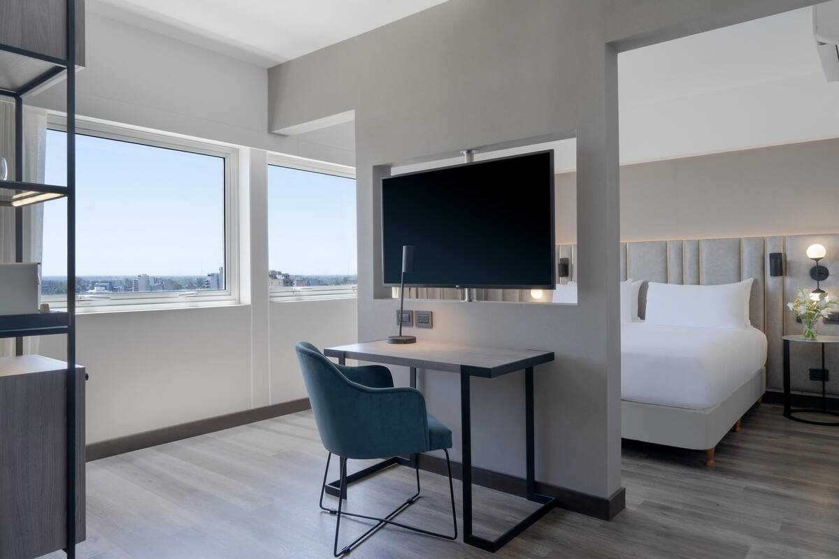 Ein modernes Hotelzimmer mit einem großen Bett, einem Schreibtisch mit Stuhl, einem Fernseher und Blick auf die Stadt durch große Fenster.
