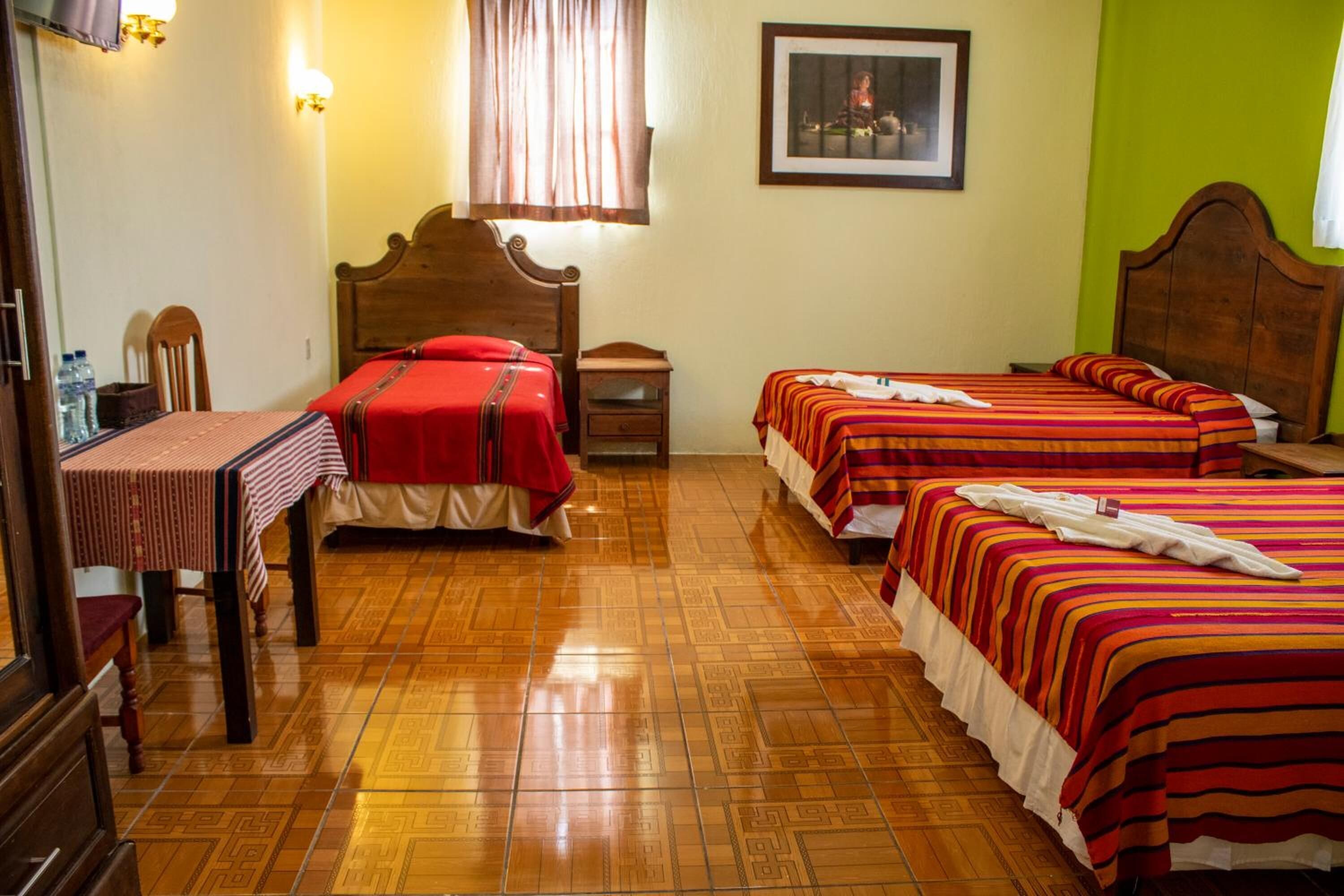 Habitación de hotel con dos camas, cabecero de madera, una mesa de comedor con mantel rojo y un cuadro en la pared.