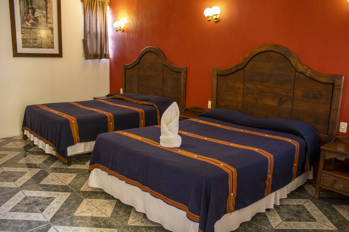 Habitación de hotel con dos camas, cada una con una colcha azul y una almohada blanca. El piso es de baldosas y el cabecero de madera.