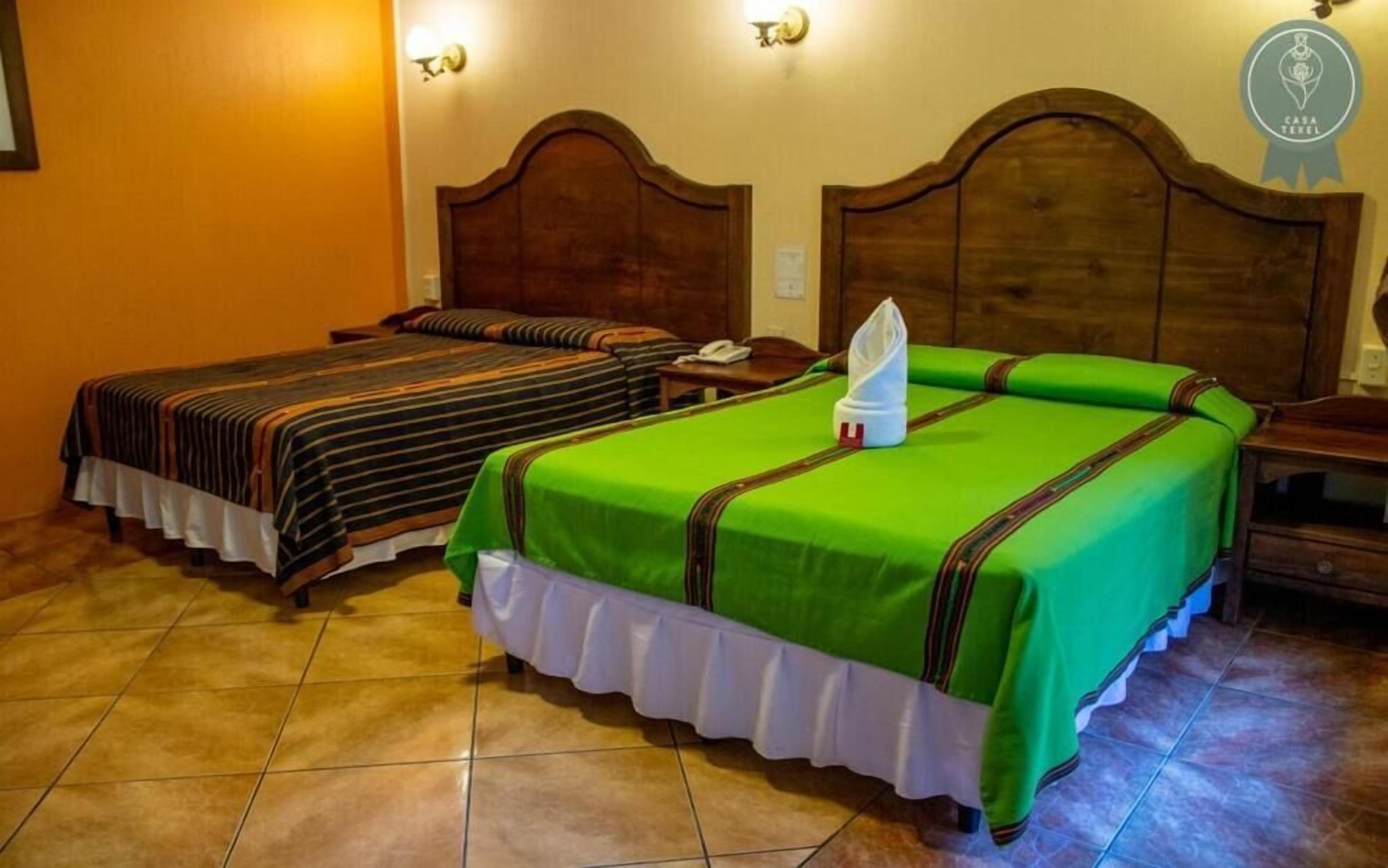 A hotel room with two beds, each with a striped bedspread and a green bedspread, wooden headboards, and a tiled floor.