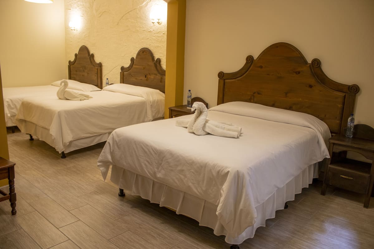 Habitación de hotel con dos camas, cada una con cabecero de madera y colcha blanca. Hay dos mesitas de noche, con lámparas y una botella de agua sobre una de ellas. El piso es de madera y la pared presenta un acabado texturizado.