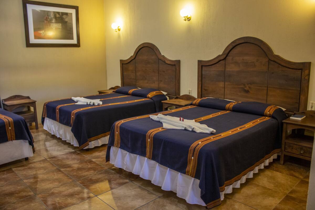 Habitación de hotel con tres camas, cada una con cubrecama azul y detalles blancos con volantes. Hay dos mesitas de noche fijadas a la pared y un cabecero de madera.