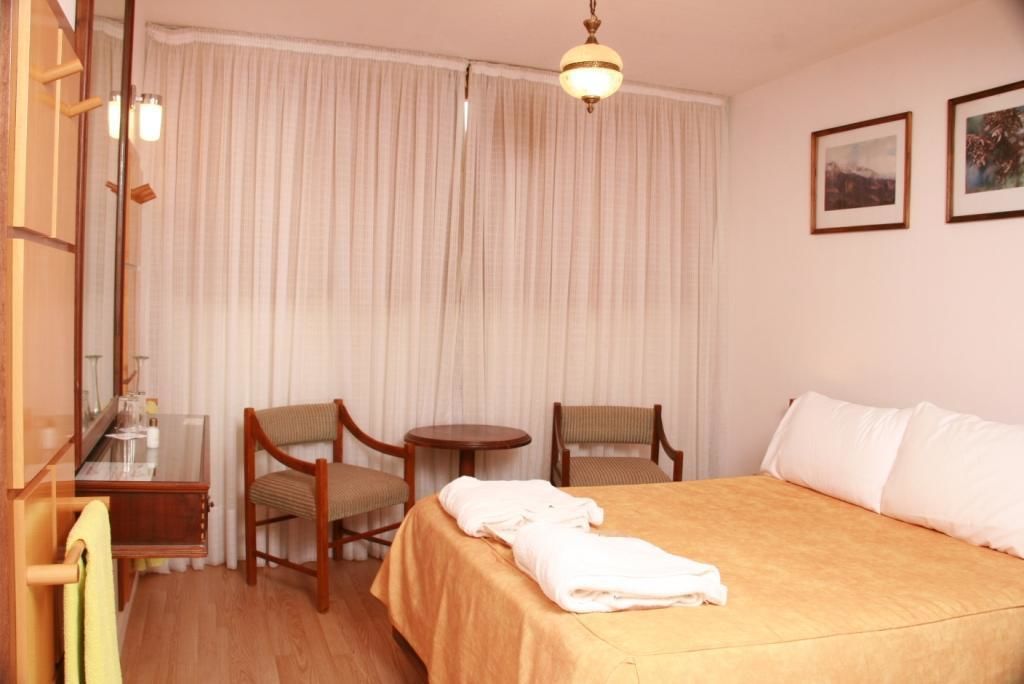 A hotel room with a bed, two chairs, a small table, a mirror, and a window with curtains.