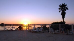 A sunset view from a terrace with lounge chairs and tables, overlooking a body of water. - Quirinale Hotel (Colon)