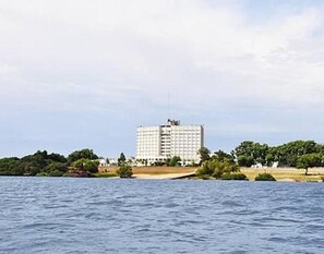 A large white building with multiple floors, situated near a body of water with greenery around.
