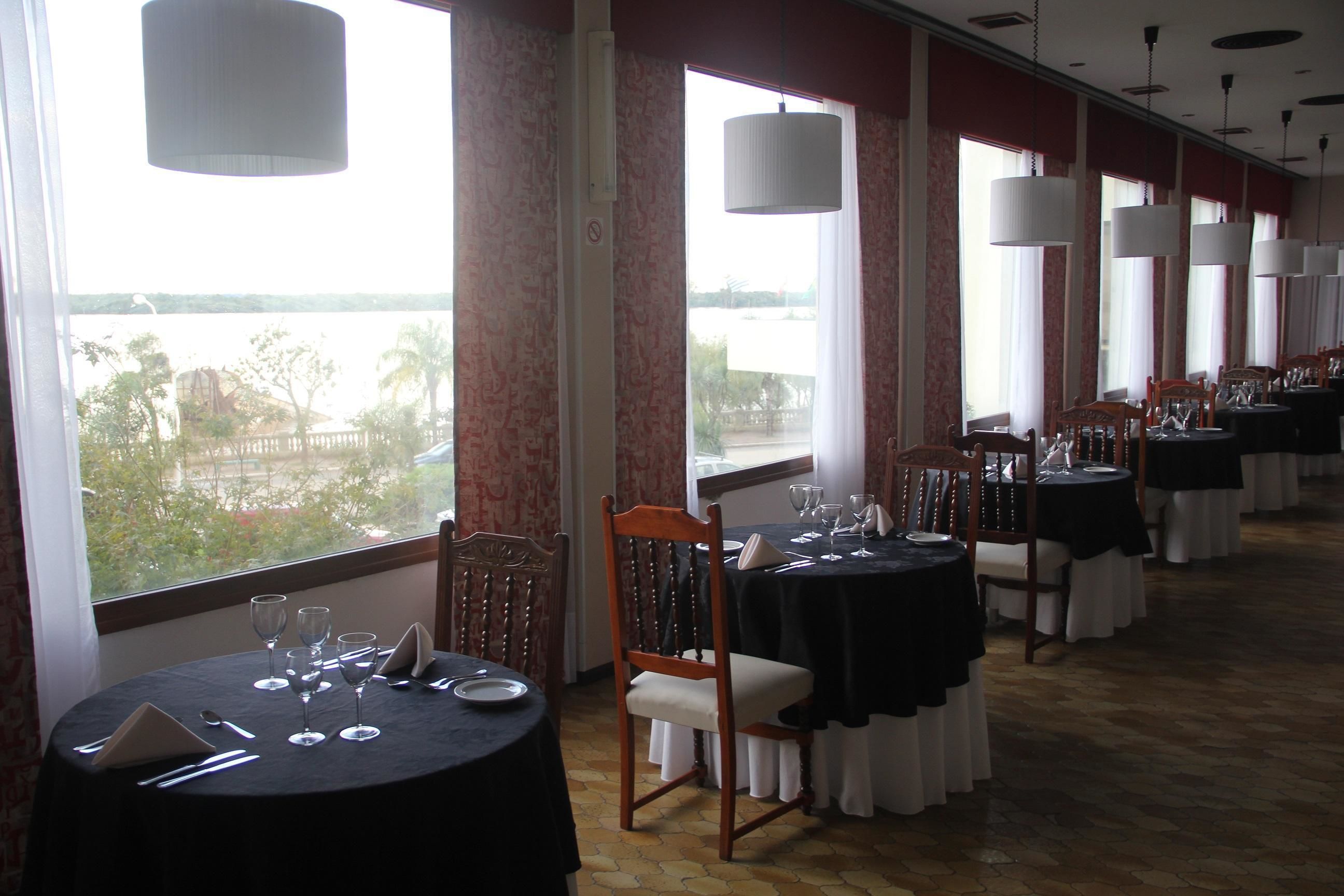 Un restaurante con mesas dispuestas para comer, amplios ventanales con cortinas e iluminación colgante.