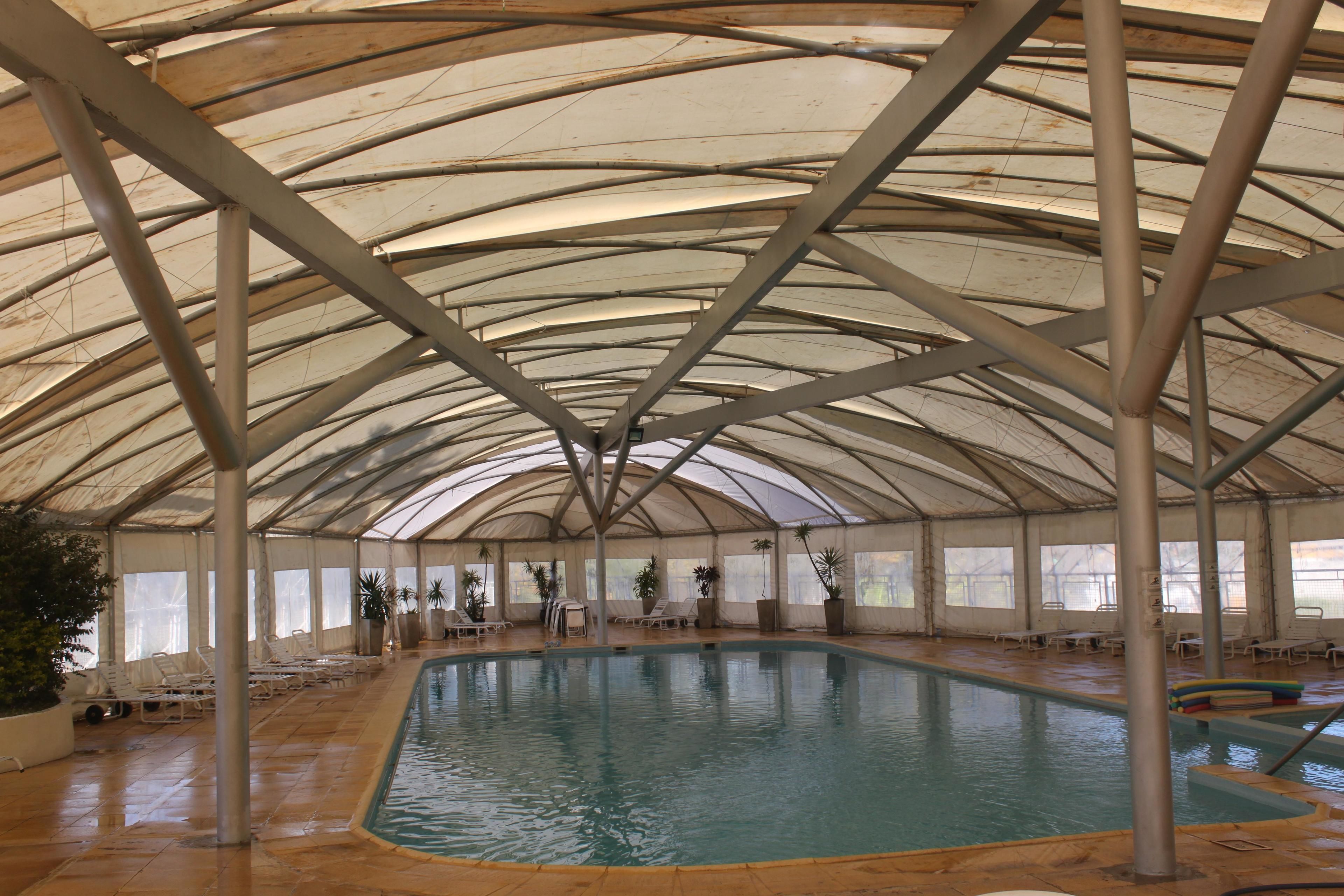 Una piscina interior con techo curvo, rodeada de sillones de descanso y plantas en macetas.