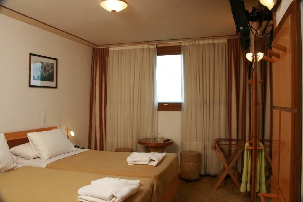 A hotel room with a bed, a small table, a chair, a waste bin, and a window with curtains.