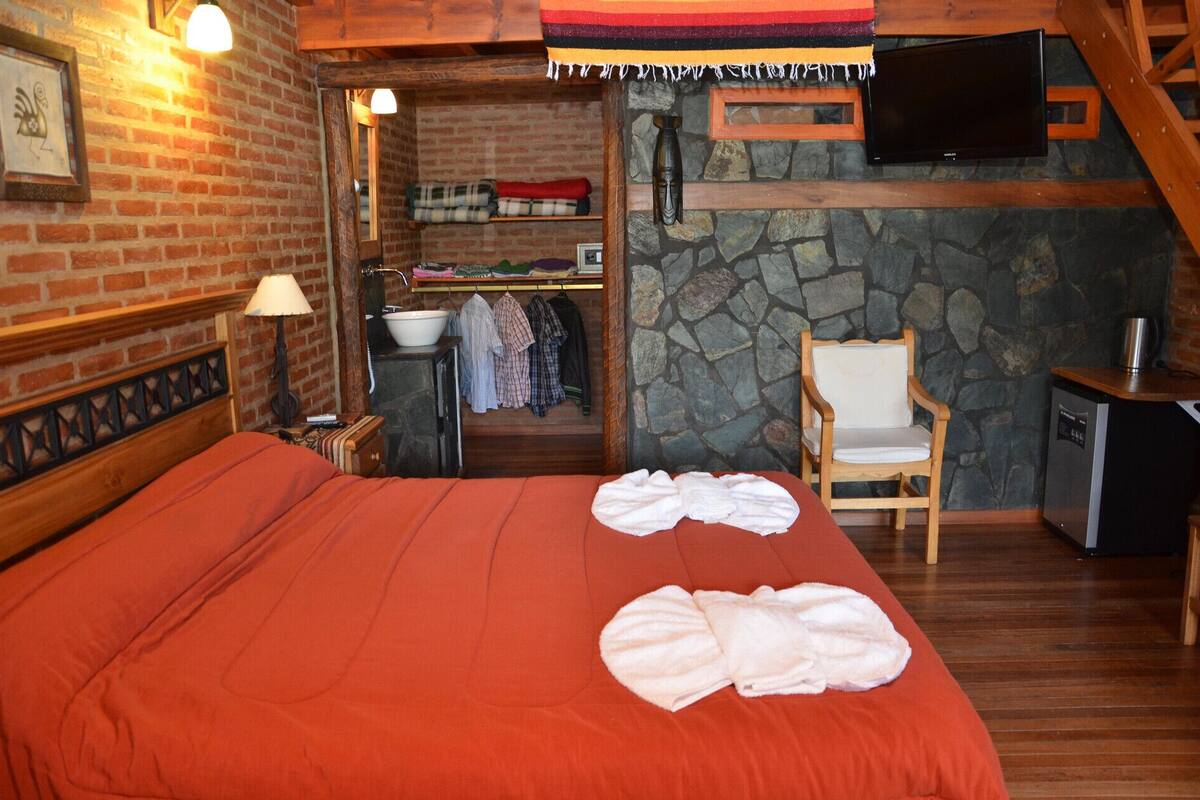A cozy bedroom with a large bed, a wooden headboard, a stone wall, a television mounted above a wooden desk, and a chair.