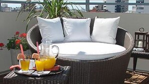 Un canapé rond en osier, agrémenté de coussins blancs, et une table en verre sur laquelle sont posées des boissons et une carafe.