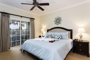 A bedroom with a bed, two bedside tables, a ceiling fan, a large window with curtains, and a decorative wall piece. - RVH 121 Centre Court Dream (Kissimmee)