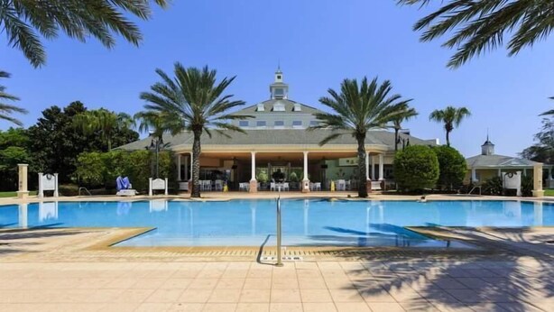 A swimming pool with a building in the background, palm trees, and a clear sky. - RVH 121 Centre Court Dream (Kissimmee)