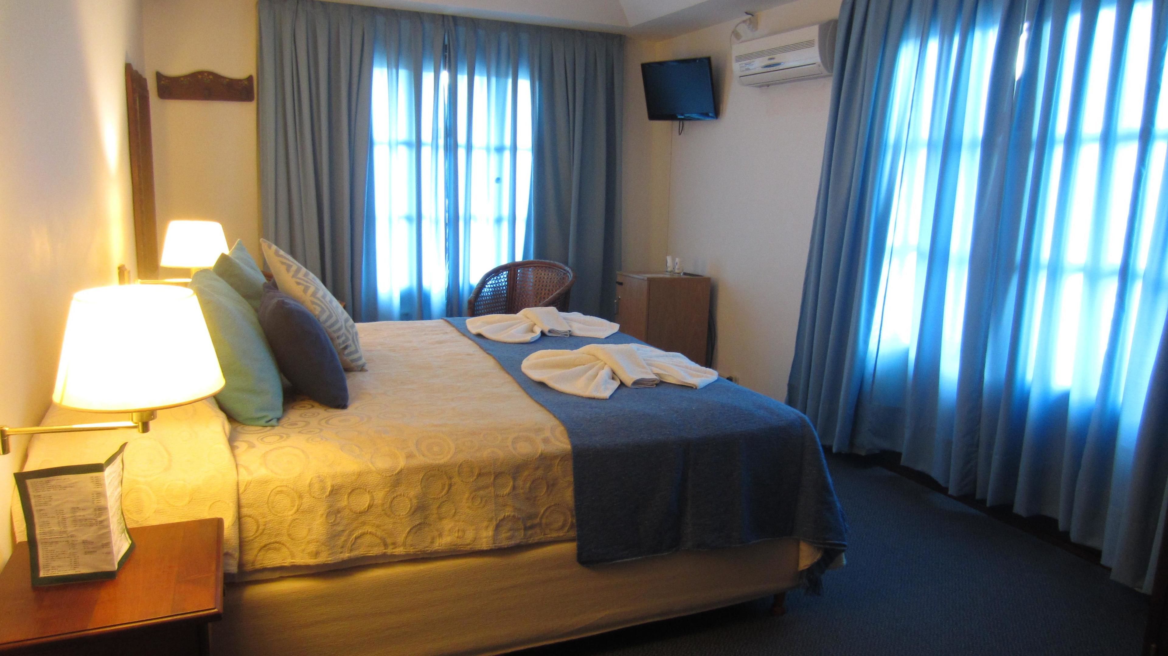 Quarto de hotel com cama, escrivaninha com luminária, cadeira, televisão e ar condicionado.