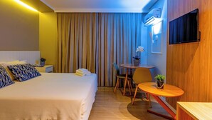 A hotel room with a bed, bedside table, lamp, curtains, a small round table with a chair, and a TV mounted on the wall. - Prive Thermas Hotel by WAM Experience (Caldas Novas)