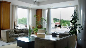 Een ruime woonkamer met een bank, een salontafel en een fauteuil. Er zijn grote ramen met gordijnen, een beeld en diverse potplanten.