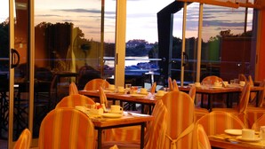Een restaurant met gedekte tafels, stoelen met gestreepte bekleding en uitzicht op een waterlichaam door grote ramen.