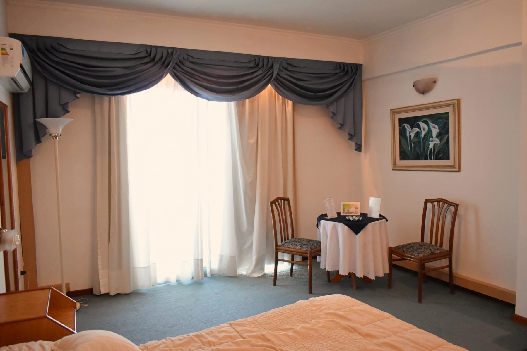 A hotel room with a bed, two chairs, a small table with a white tablecloth, a painting on the wall, and a large window with curtains.