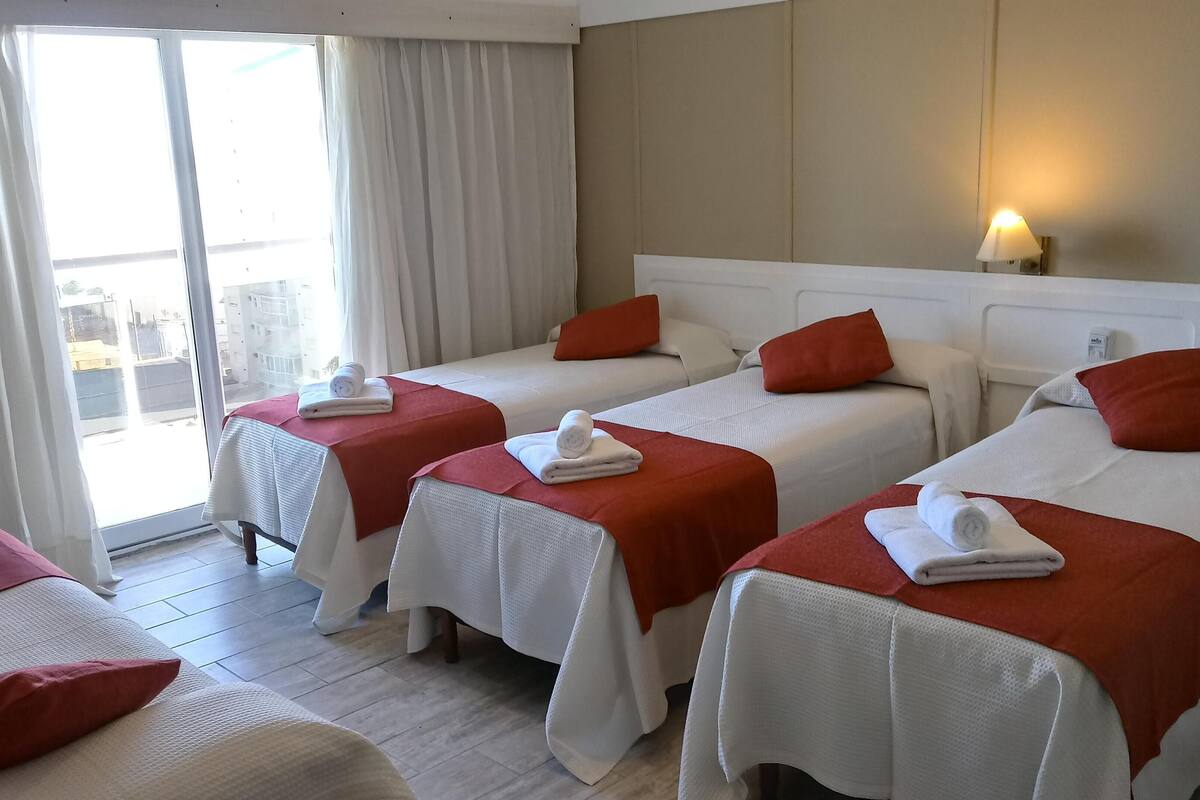 Ein Hotelzimmer mit zwei Einzelbetten, jeweils mit einer roten Decke und einem zusammengerollten Handtuch. Es gibt ein großes Fenster mit Vorhängen, einen Nachttisch mit einer Lampe und einen Blick auf Gebäude im Freien.