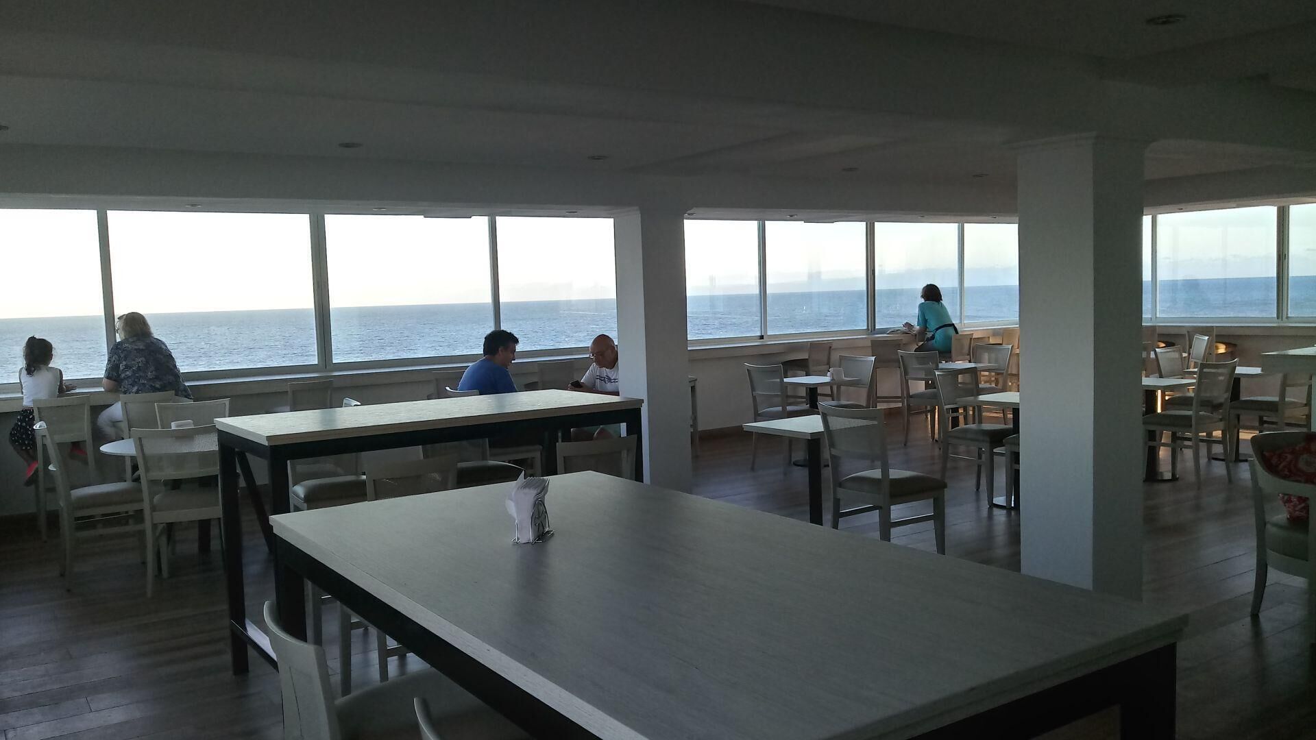 A restaurant with large windows offering a view of the sea. Tables and chairs are arranged for dining.