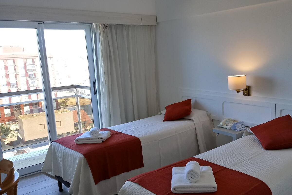 Ein Hotelzimmer mit zwei Betten, jedes mit einem roten Kopfkissen und einem weißen Handtuch. Ein Nachttisch mit Lampe und Telefon ist vorhanden. Das Zimmer verfügt über ein großes Fenster mit Vorhängen und einen Blick auf Gebäude.