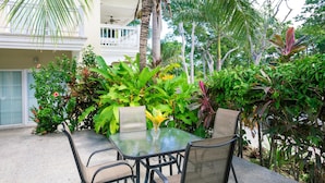 Un patio al aire libre con una mesa y sillas de vidrio, rodeado de exuberante follaje tropical y una palmera.