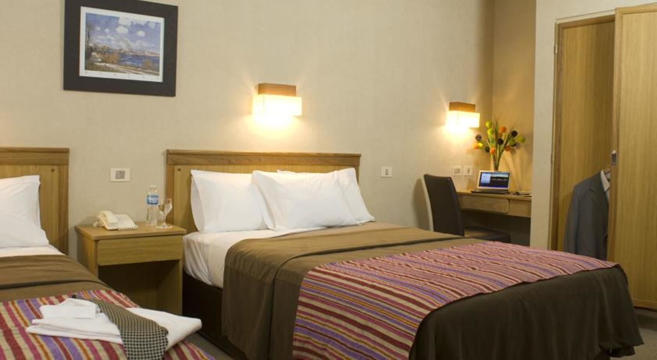 Habitación de hotel con dos camas, un escritorio con una computadora portátil, una silla, una mesita de noche con un teléfono y gafas, y un cuadro en la pared.