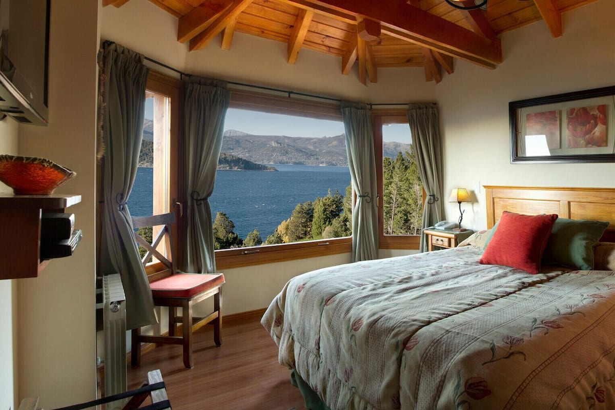 A bedroom with a large bed, a wooden headboard, a nightstand with a lamp, a chair, and a window with curtains.