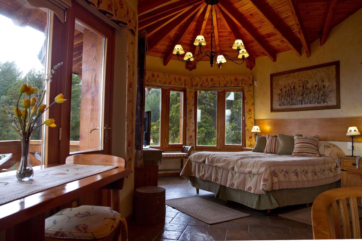 A cozy room with a bed, a desk, and a chair, featuring large windows and a wooden ceiling.