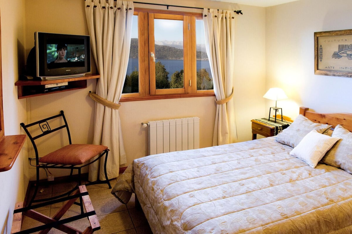 A bedroom with a bed, a chair, a television, a window with curtains, a radiator, and a wall-mounted picture.