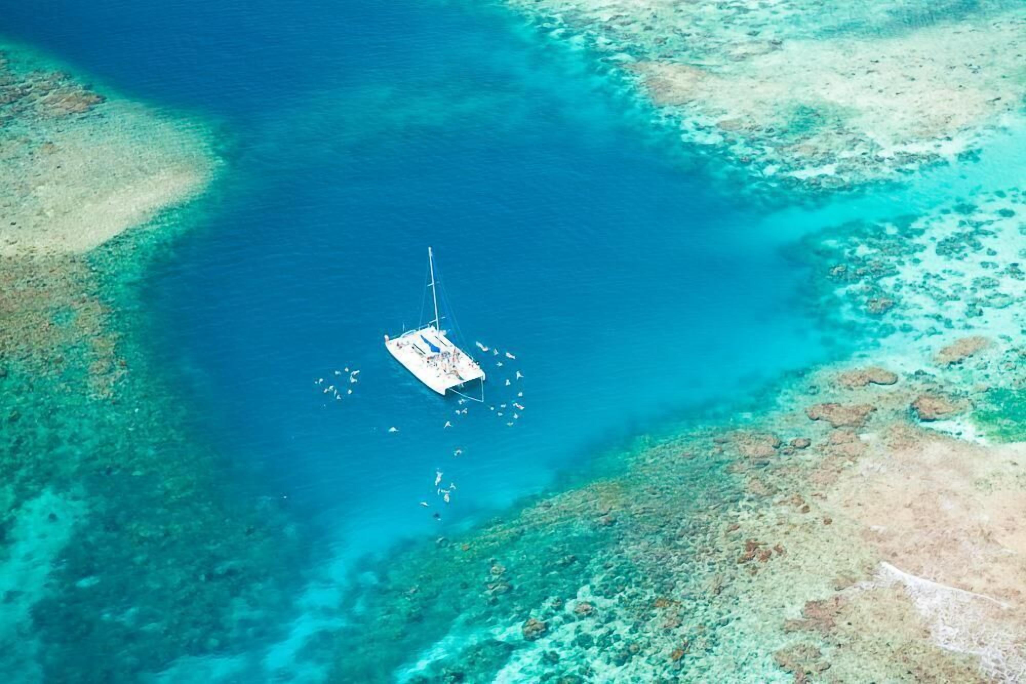 An aerial view of a boat anchored in clear blue waters with coral reefs nearby.