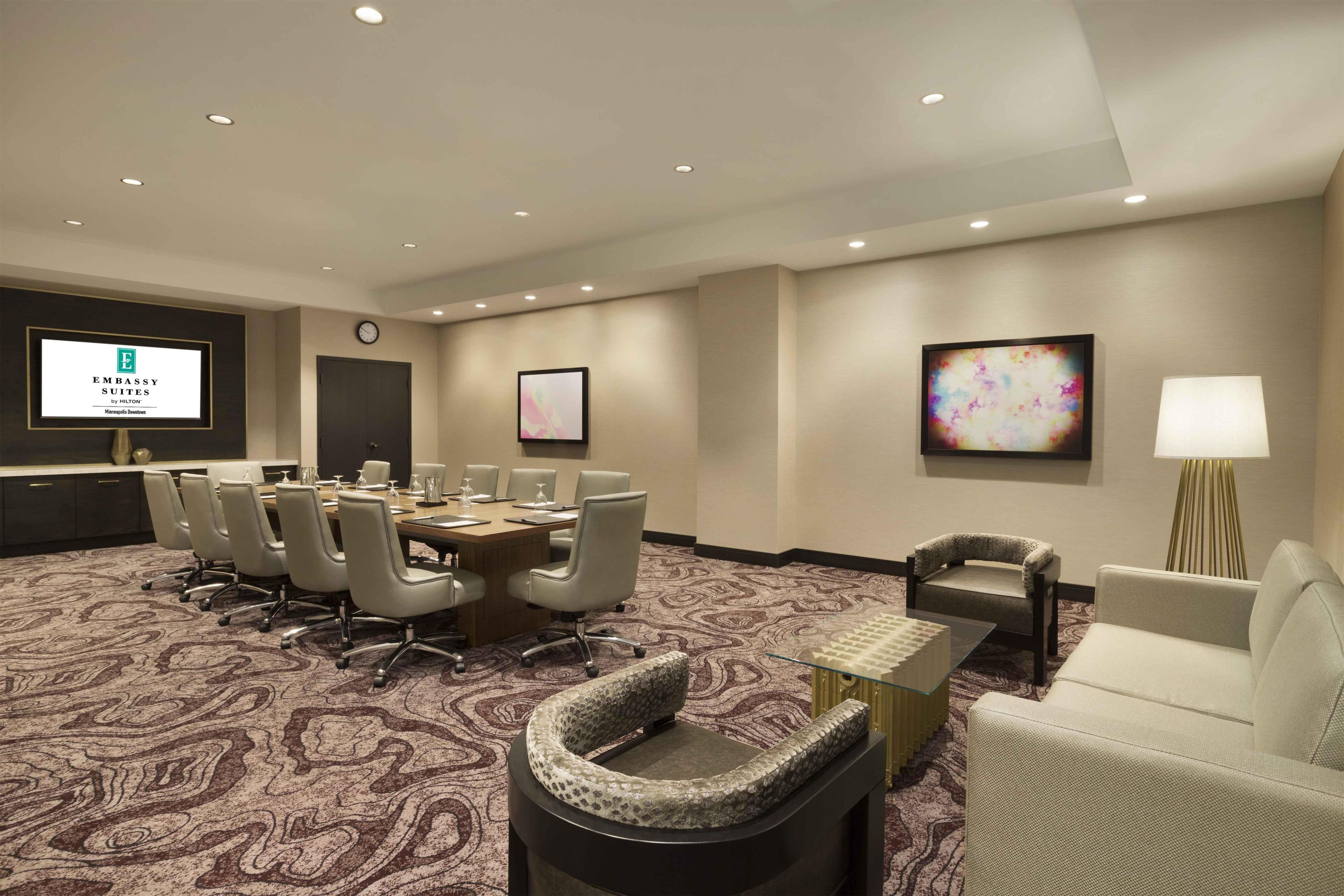 A conference room with a long table, chairs, a clock, and a screen displaying 'Embassy Suites by Hilton Minneapolis Downtown'.
