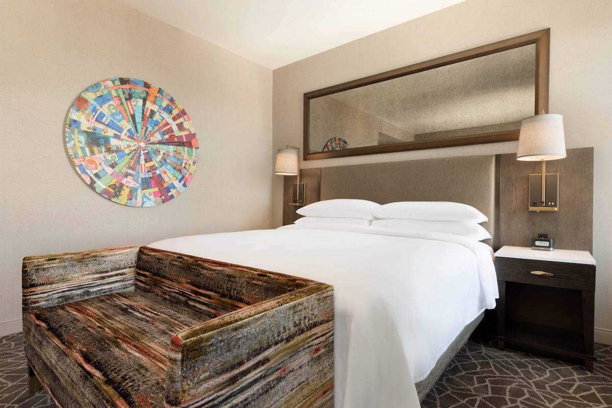 Habitació d'hotel amb un llit, una obra d'art rodona penjada a la paret, una banca de fusta, una tauleta de nit amb una làmpada i un mirall sobre el llit.