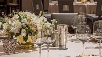 A dining table set with white tablecloths, clear glassware, and a centerpiece of white flowers with gold accents.