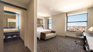 A hotel room with a bed, a desk, a chair, a large window, and a bathroom with a mirror and sink.