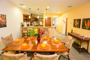 A dining area with a wooden table set for six, surrounded by wicker chairs. A kitchen is visible in the background.
