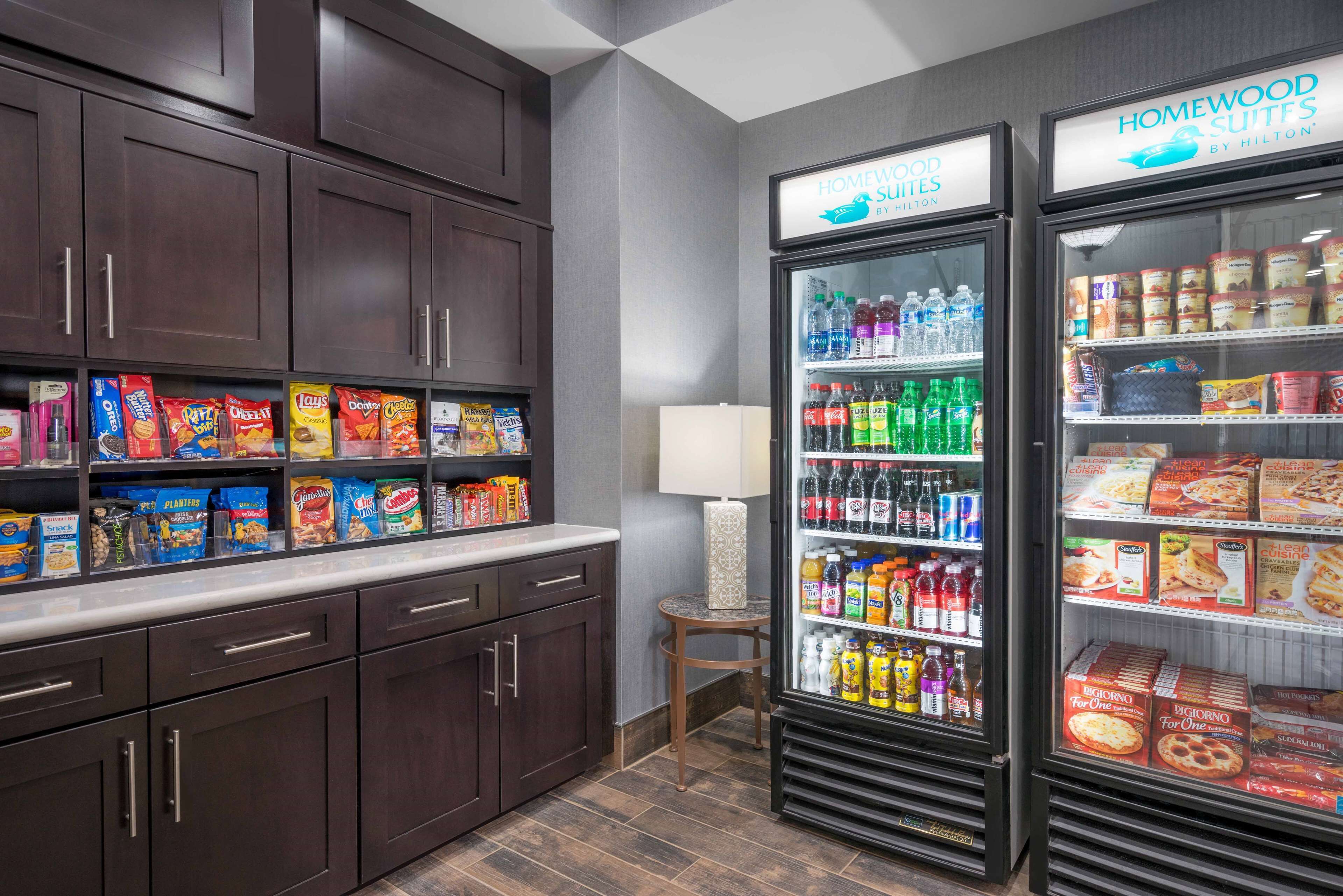 A kitchenette with dark wooden cabinets, a countertop with snacks, and two beverage refrigerators labeled "Homewood Suites by Hilton".