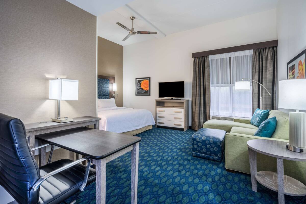 A hotel room with a bed, a desk with a chair, a sofa, a TV, and a window with curtains.