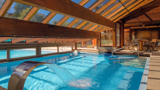 Una piscina con techo de cristal, agua azul cristalina y sillas de madera en la terraza.