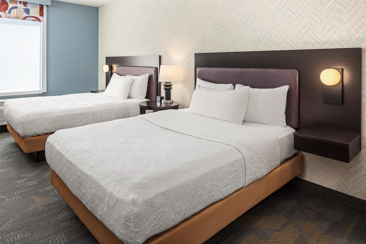 A hotel room with two beds, each with a headboard and bedside table, and a large window.