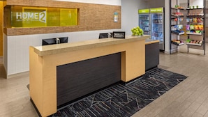A hotel reception area with a reception desk, a 'Home 2 Suites by Hilton' sign, a 'Hilton' sign, a 'COLD' and 'Really Cold' beverage refrigerator, and a snack shelf. - Home2 Suites by Hilton Roseville Minneapolis (Roseville)