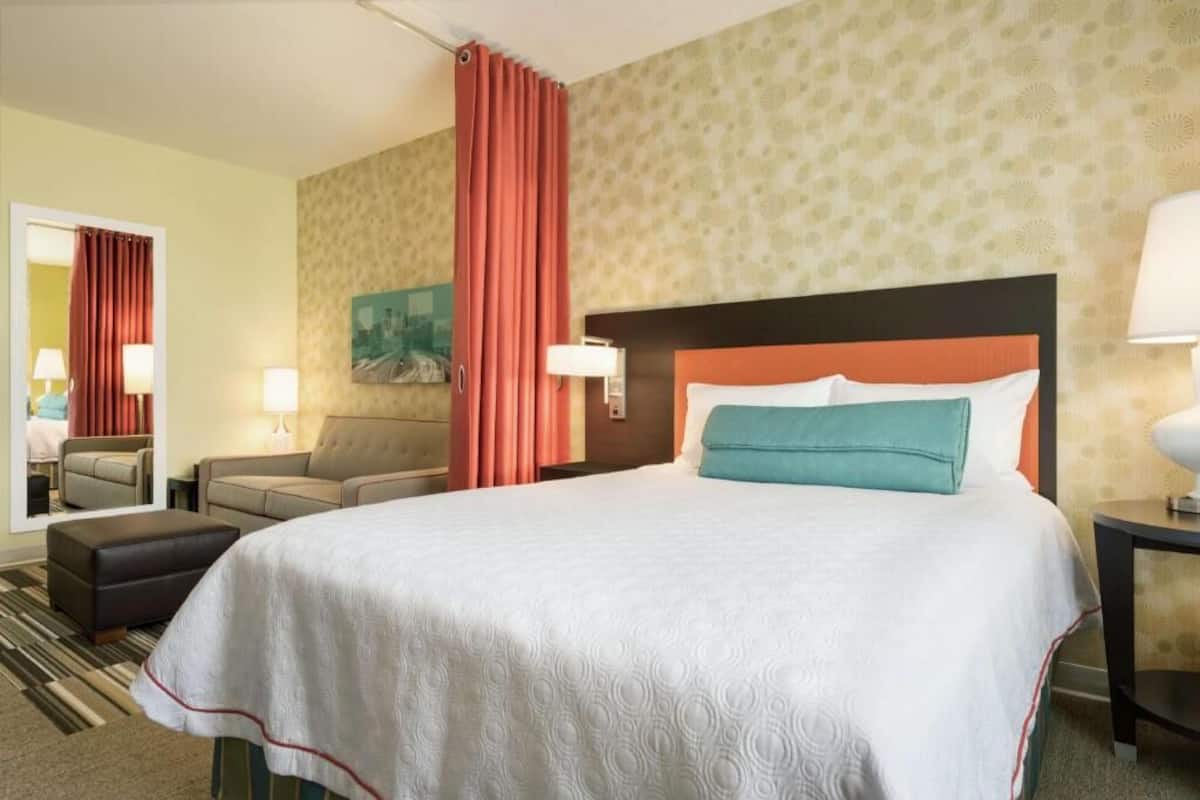A hotel room with a bed, a sofa, a nightstand, a lamp, and a mirror.