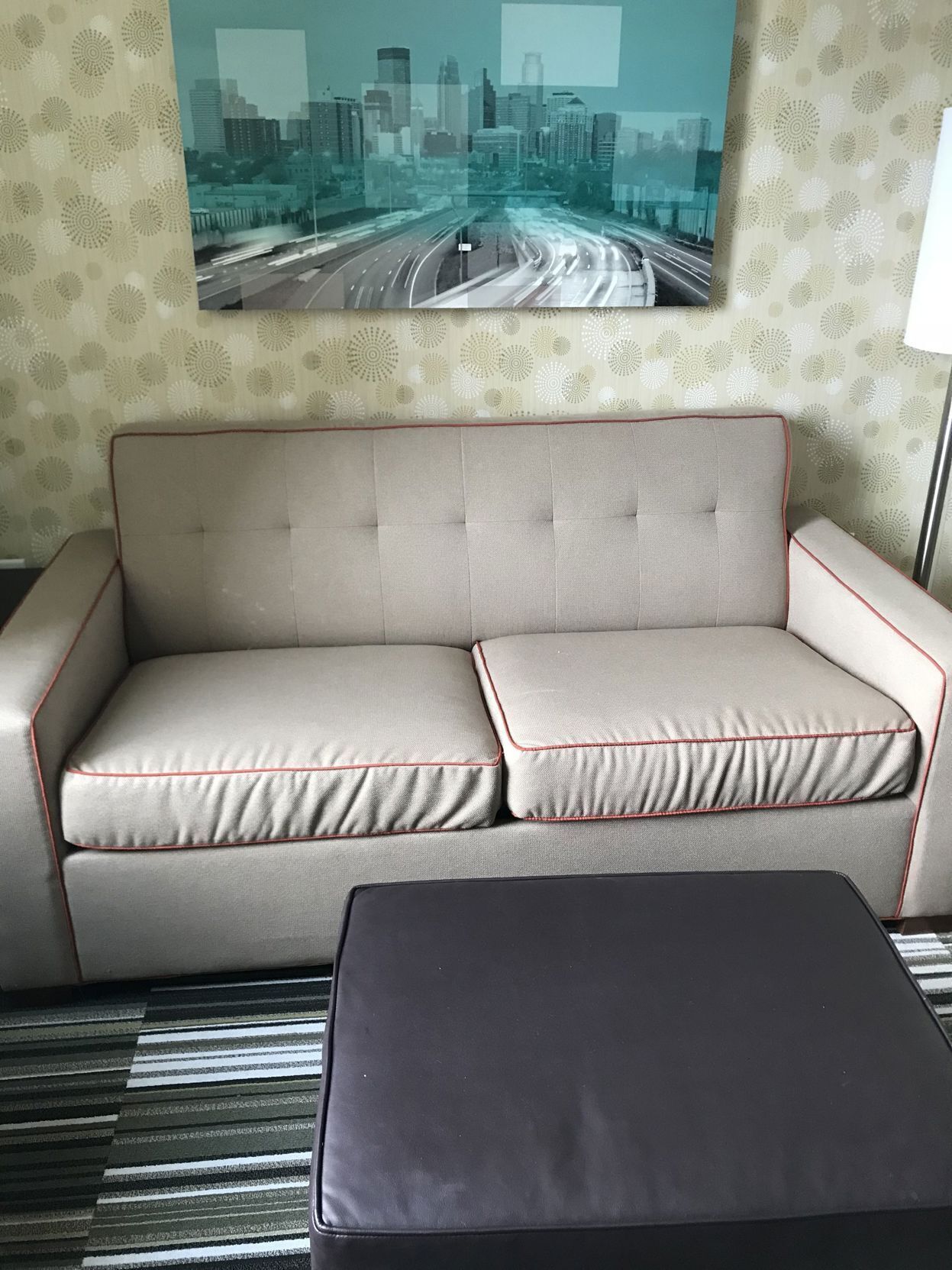 A beige sofa with red piping, a black ottoman, and a cityscape view through a window.